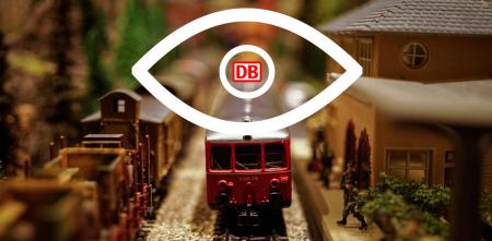 Ein großes Auge, dessen Pupille aus dem Logo der Deutschen Bahn AG besteht, überblickt einen Miniaturbahnhof.