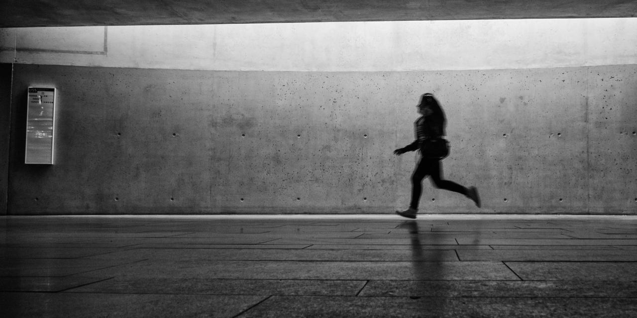 Eine Person, die, an einer Bahnstation, von rechts nach links rennt (Aufnahme in schwarz-weiß).
